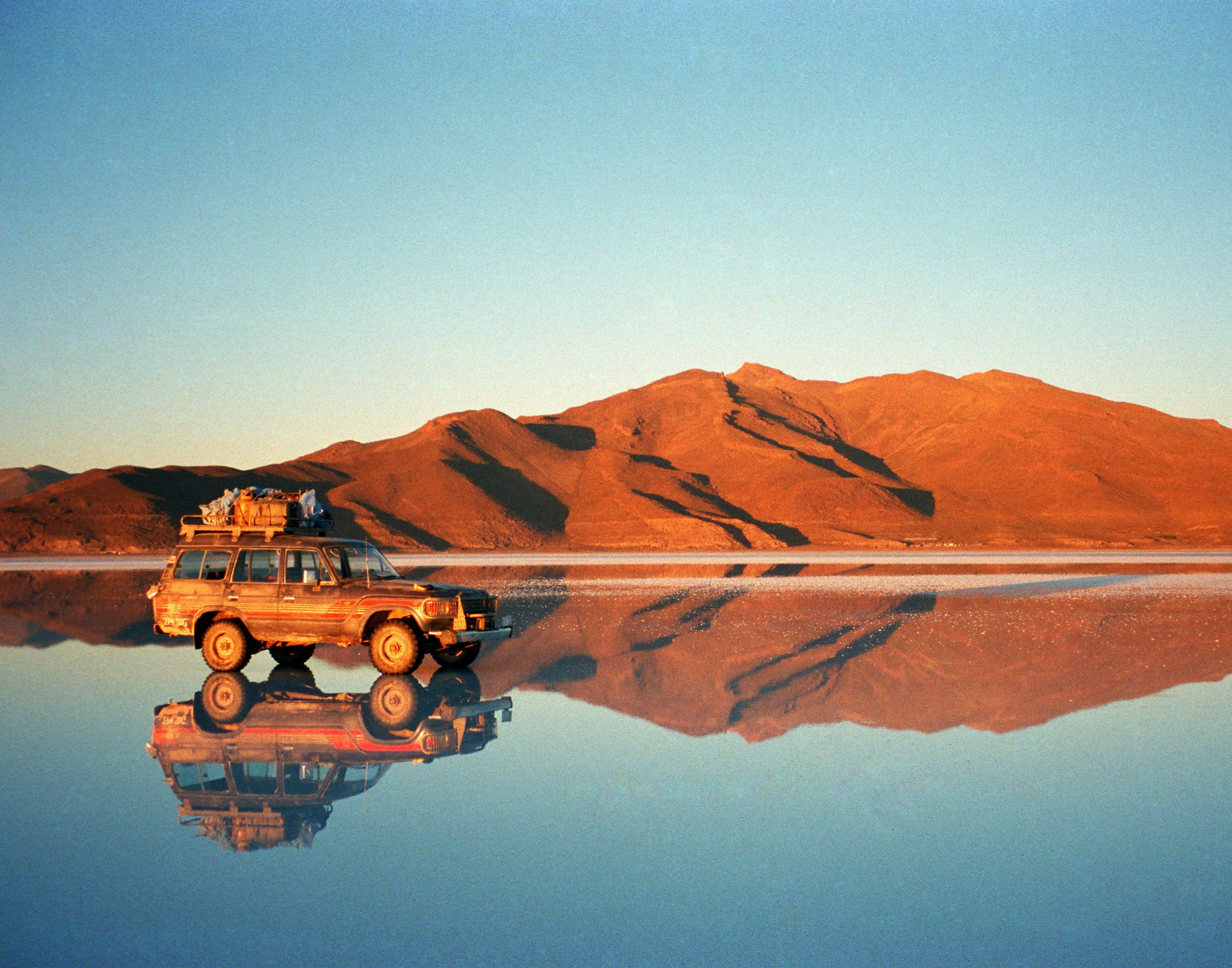 4x4 chargé roulant sur le miroir du salar d'Uyuni au soleil couchant, montagnes ocre reflétées