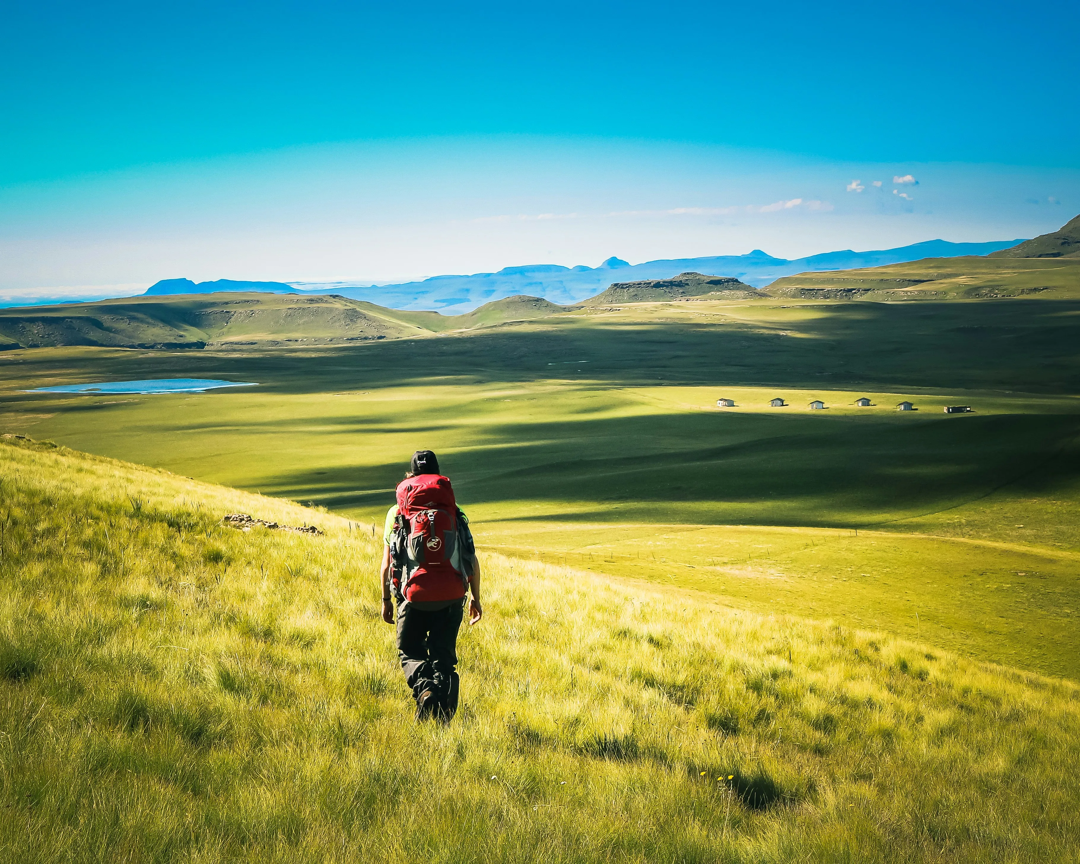 Randonneur au sac rouge descendant vers un lac des hauts plateaux verts du Lesotho