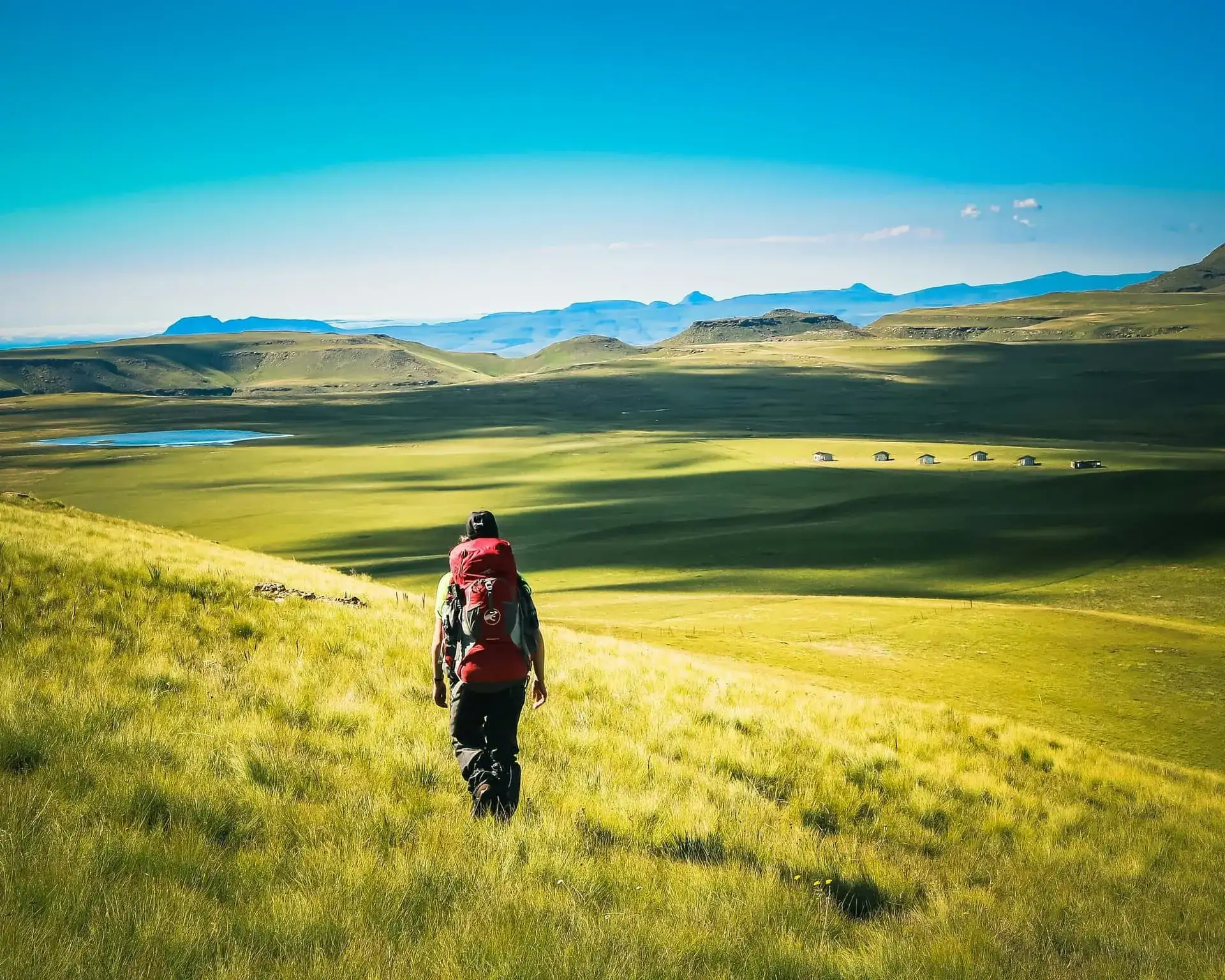 A hiker with a red pack descending towards a lake on the green high plateaus of Lesotho