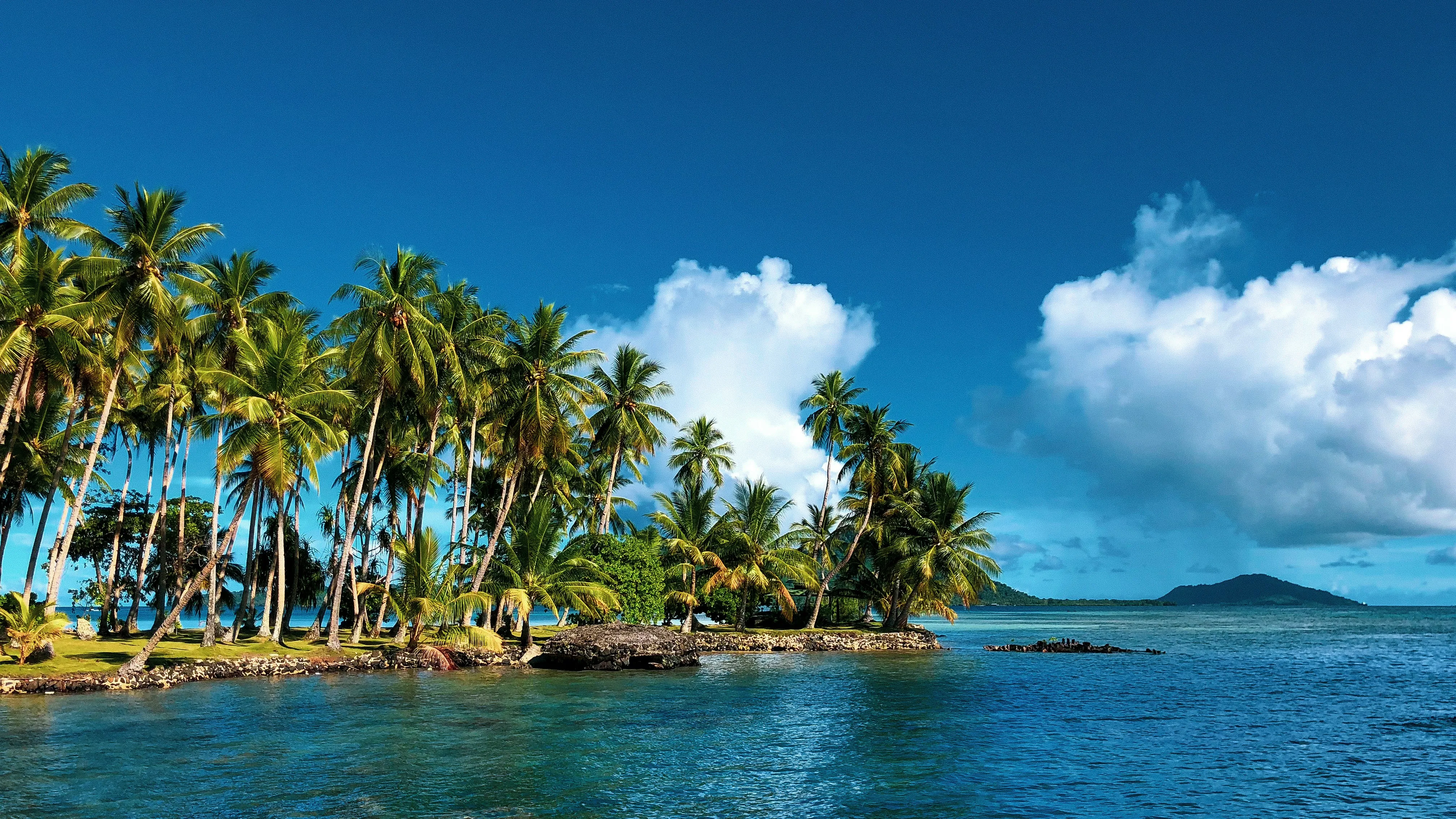 Îlot de cocotiers penchés sur un lagon turquoise de Micronésie, île volcanique à l'horizon